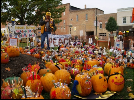 A man in a city square or common area looks at a large number of Jack-o-lanterns, most of which are brightly painted.
